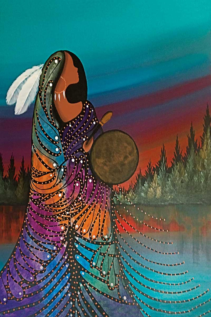 A woman in a colourful gown and white feathers in her hair. She holds a leather drum. She stands at the edge of a lake, trees surrounding it.