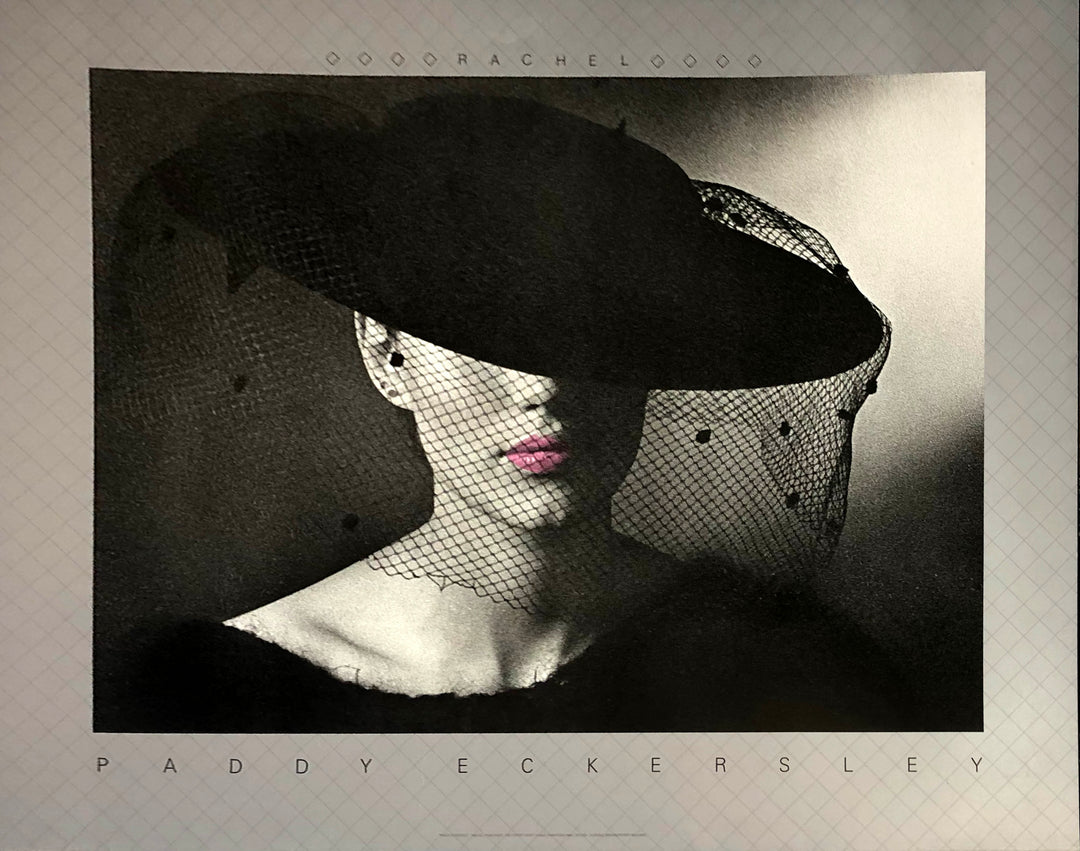 black and white photo. A pale woman wears a black dress and hat, her collar bone up only visible. A veil falls from the wide brim hat, which covers her eyes. Her lips are in colour. a pink.