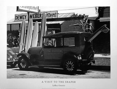 Black and white photo. A dark, vintage car is parked by the curb. Near by are stood up surfboards and a sign behind them reading "Dewey Weber's Surfboards"