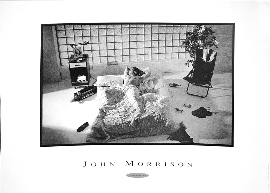 a black and white photo. A man lies in his bed which sits in the middle of the room, Clothes are strewn across the floor, with heels and a top among them. There is also a wine bottle a record player on the floor. A speaker with the picture of a woman and lamp on it stands near the record player. A folding chair and plant are on the other side of the bed. Cubic glass is in the wall behind the bed. A light comes in from the left.
