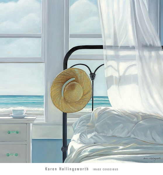 A straw hat rests on the black, metal bed frame of a white bed. A white bedside table with a teacup and saucer sits next to the bed. A white curtains blows onto the bed. The open windows overlook a beach, waves rolling onto the shore.