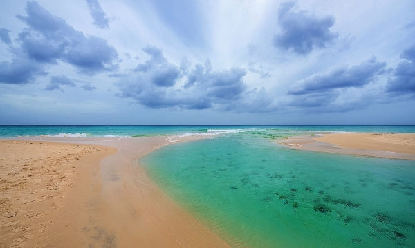 A scenic aerial view of a tropical beach with pale sand, turquoise water, and a dramatic cloudy sky.