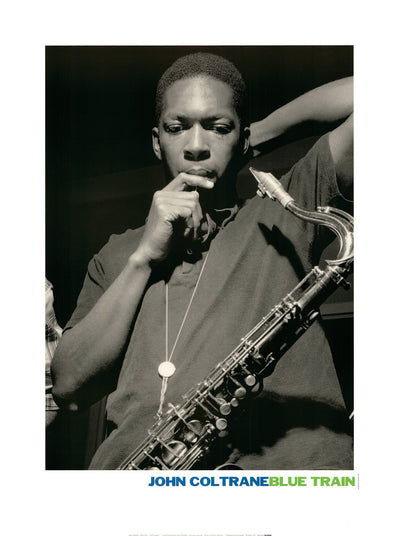 A black and white photo of John Coltrane during his recording of his album Blue Train. He has his saxaphone on him, holding a finger across his lip. His other hand is behind his head.