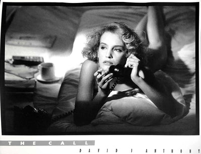 A blonde, caucasian woman lays on her bed, chest on the sheets. She rests on a pillow, smoking a cigarette with a black phone in her hand.