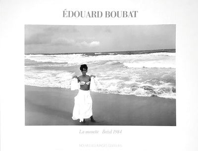 black and white image. A woman in a white dress takes off her white shirt to reveal her white bra. She stands in the sand next to crashing waves. 