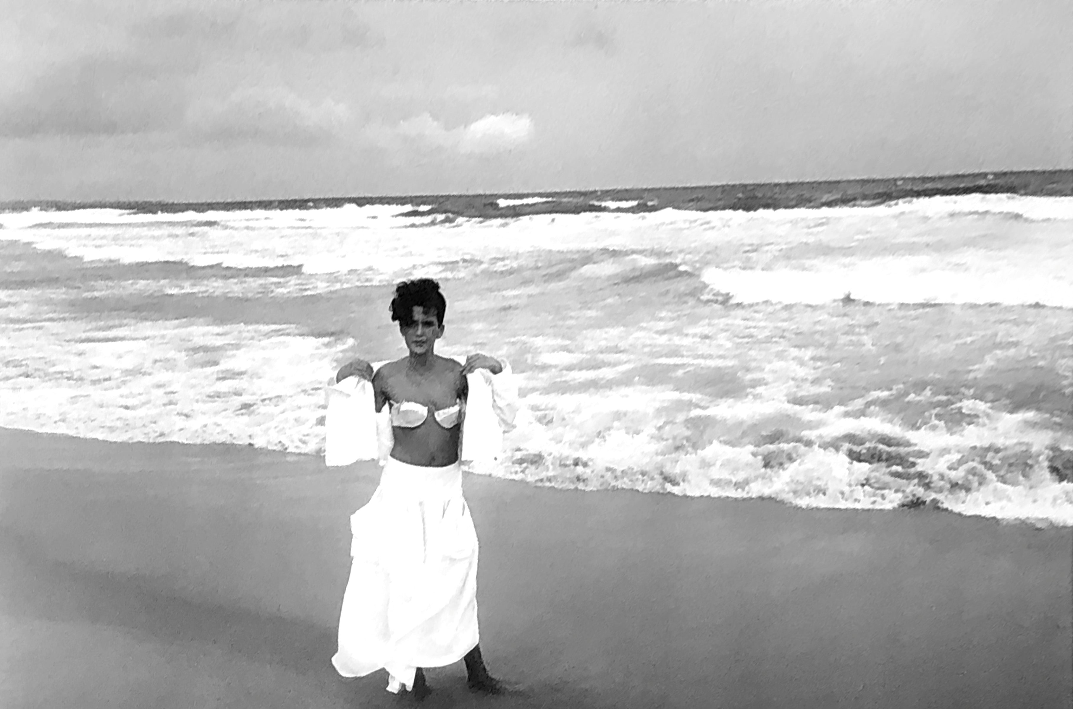 black and white image. A woman in a white dress takes off her white shirt to reveal her white bra. She stands in the sand next to crashing waves. 