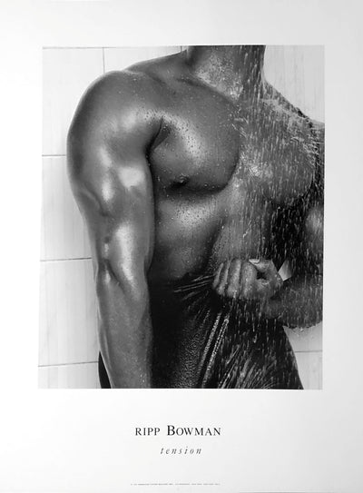 A naked chest and arm, both fairly muscle bound. The model is sprayed with water like a shower. They hold their trunks in their other hand. The model has a white tile wall behind them.