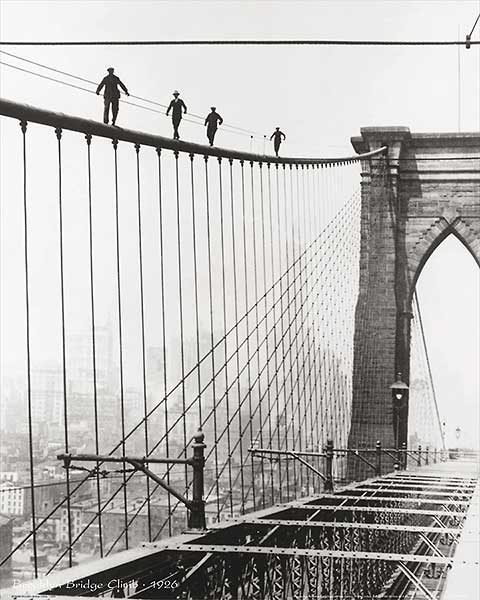 Black and white photo. Men walk along the suspension wire of the Brooklyn Bridge