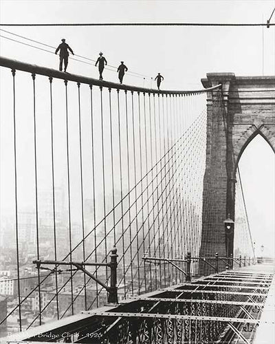 Black and white photo. Men walk along the suspension wire of the Brooklyn Bridge