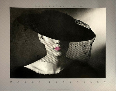 black and white photo. A pale woman wears a black dress and hat, her collar bone up only visible. A veil falls from the wide brim hat, which covers her eyes. Her lips are in colour. a pink. 