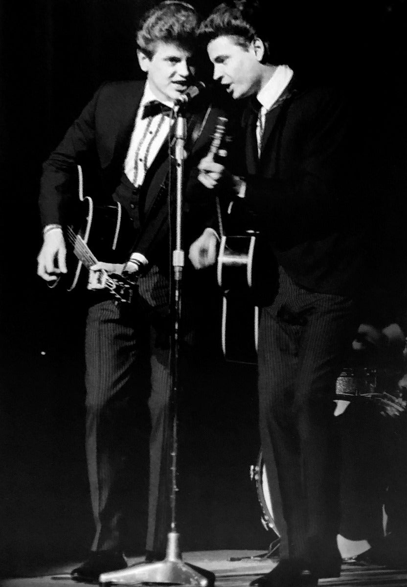 Black and white photo of two men in suits performing on stage, each holding an acoustic guitar, with a microphone stand in front of them.