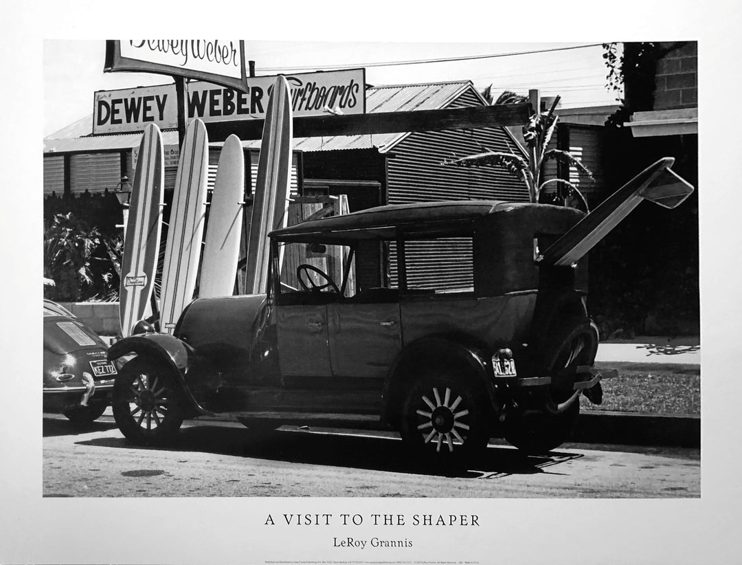 Black and white photo. A dark, vintage car is parked by the curb. Near by are stood up surfboards and a sign behind them reading "Dewey Weber's Surfboards"