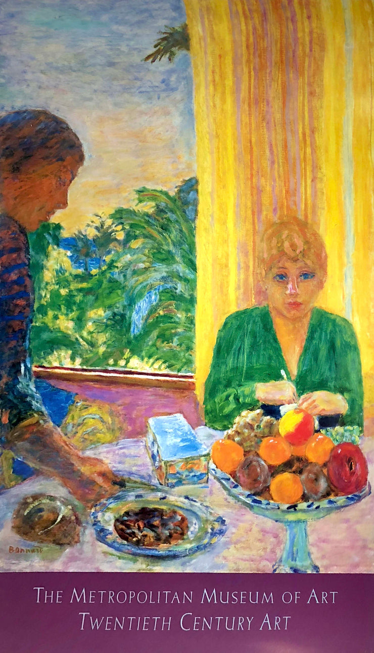  A woman in a green blouse sits in front of a yellow curtain. She is at a table covered with food. Someone comes by and places more food on the table. Lush bushes sit outside the window. 