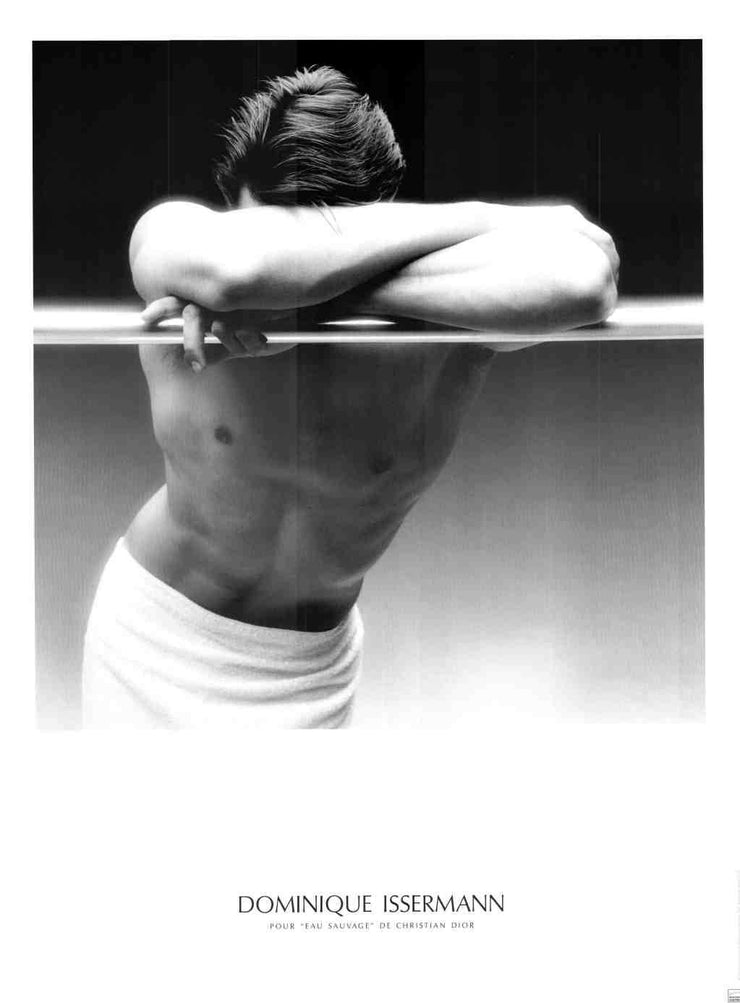A black and white photo of a buff, shirtless man. He rests his muscular arms on a horizontal, metal bar. His arms hide his face. He wears a white loincloth which shows off his chest.