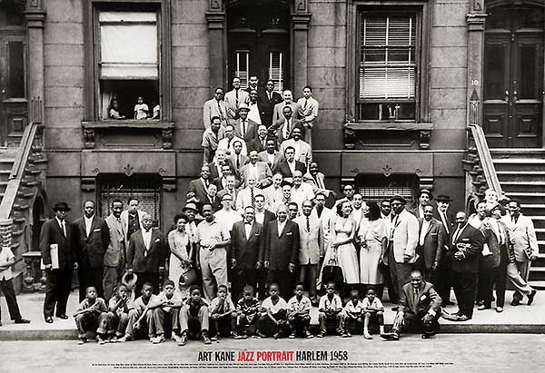 black and white photo. A group photograph of several people outside a house in Harlem. The people are dressed in black and white suits. Several stand on the steps up to the building, while others line the sidewalk. Children sit along the sidewalk. Text reads: Art Kane, Jazz Portrait [in red], Harlem 1958." end of text.