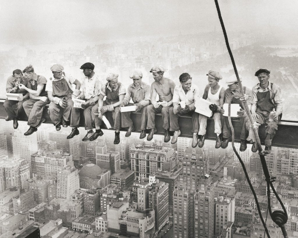 A vintage black and white photo of eleven construction workers having their lunch on a support beam with the city (New York City) laying underneath them. They wear overalls and flat caps.