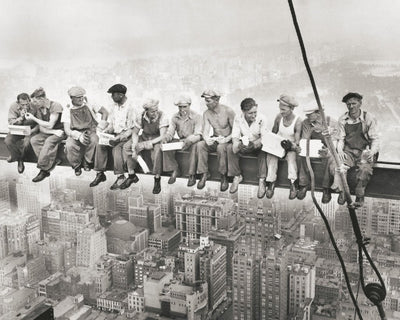 A vintage black and white photo of eleven construction workers having their lunch on a support beam with the city (New York City) laying underneath them. They wear overalls and flat caps.