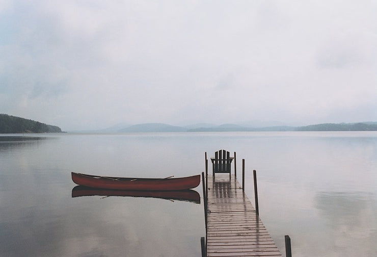 A lone. red canoe on a calm lake. It is floating next to a plank dock with a muskoka chair at the end of it. The sky is overcast, turning the water a silver grey.