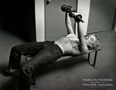 A black and white photo of Marilyn Monroe lifting weights. She lays on a bench in jeans and a sports bra, lifting the weights above her.