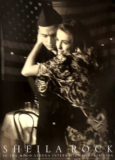 A slightly blurry image of a woman in a floral dress dancing with a soldier in a dark uniform. There is a table and chair in the background. An american flag hangs in the background.