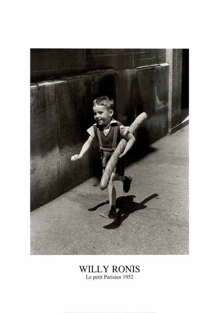 A black and white photo print of a young boy running down the road/sidewalk with a full baguette, which is nearly as big as he is. 