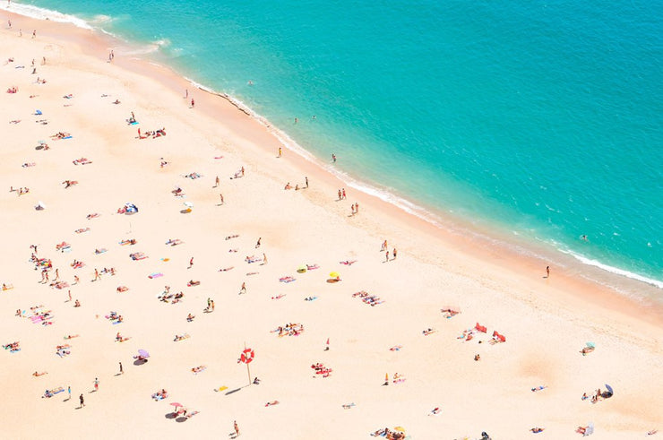 An aerial photograph of a populated beach. The pale sand has umbrellas and people scattered around like ants. The sea is a vibrant turquoise.