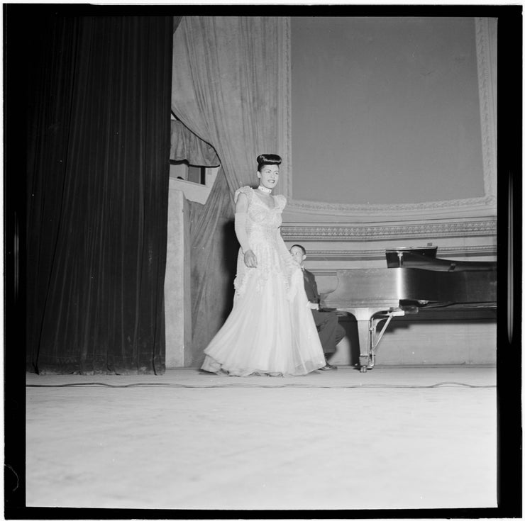 Black and white portrait of Billie Holiday at Carnegie Hall in New York City, N.Y. (1947). She walks out onto the stage in a white gown past the pianist and his piano.