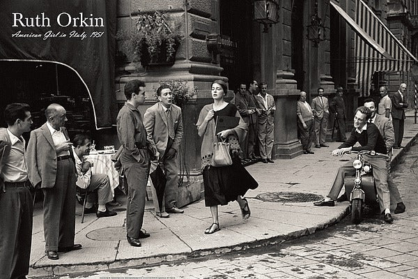 Black and White photo. A young, caucasian woman walks along a sidewalk to a corner. Several men are looking at her. One sits on a moped, smiling at her. Another whistles or is making kissing lips at her. She appears to be in distress. 