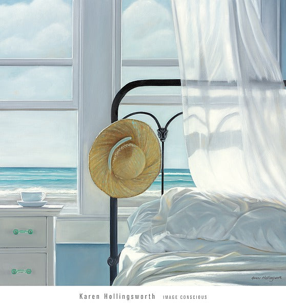 A straw hat rests on the black, metal bed frame of a white bed. A white bedside table with a teacup and saucer sits next to the bed. A white curtains blows onto the bed. The open windows overlook a beach, waves rolling onto the shore. 