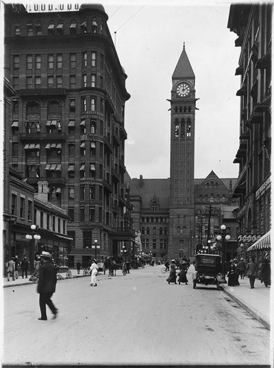 A black and white photo of Toronto's Old City Hall from Bay Street. People can be seen crossing the street. An automobile is parked at the side of the road.